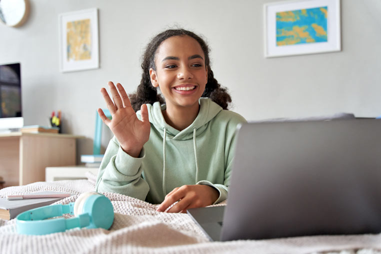 A smiling teenage girl waves to the camera during a video call  in her bedroom.
